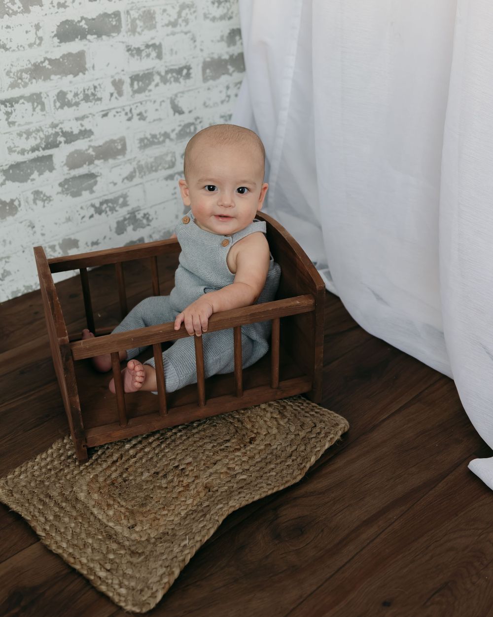 A baby in blue sitting in a vintage wooden rocking cradle on a jute rug.