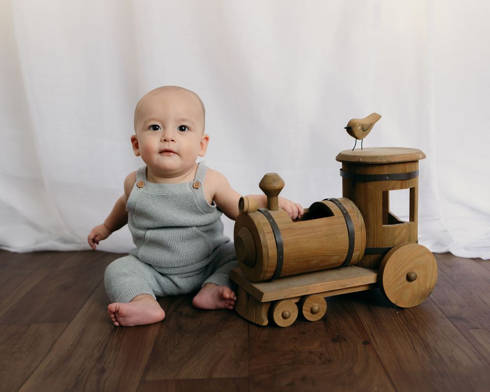 A baby in gray sits beside a wooden toy train on hardwood flooring.