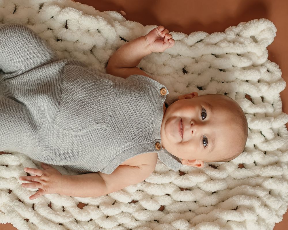 A tiny baby wearing gray rests on a chunky knit cream-colored blanket.
