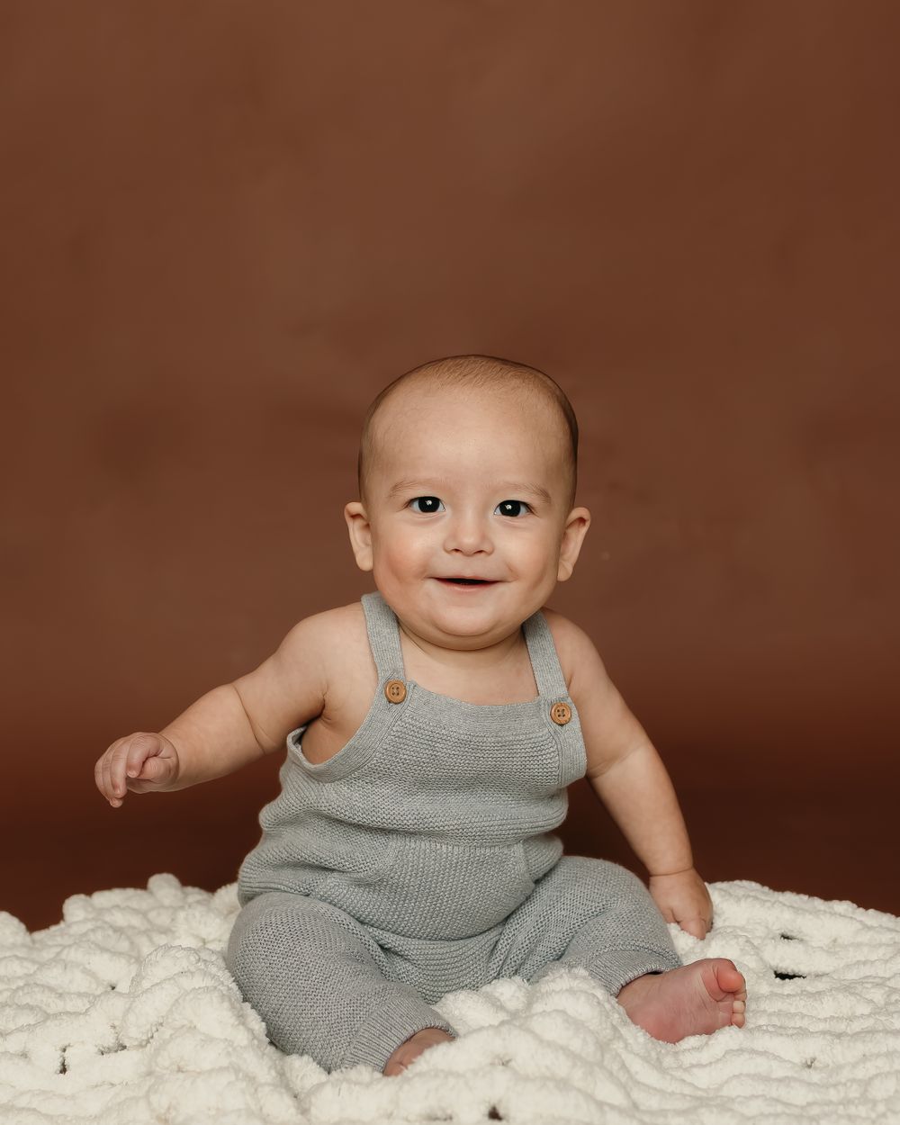 Baby in gray overalls sits on white textured fabric against brown background.
