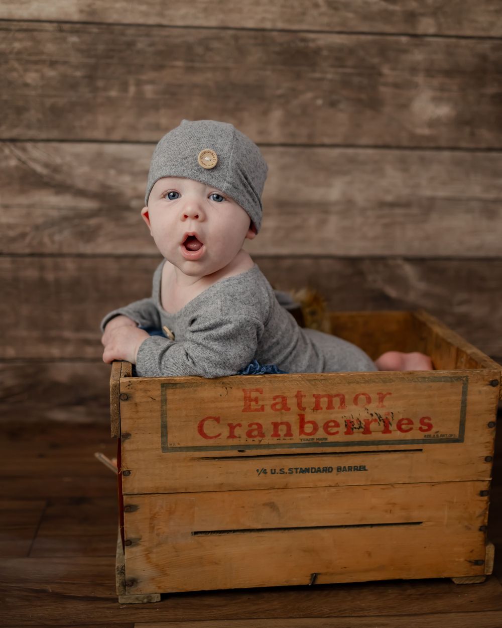 Baby in gray outfit and cap poses in vintage Eatmor Cranberries wooden crate.