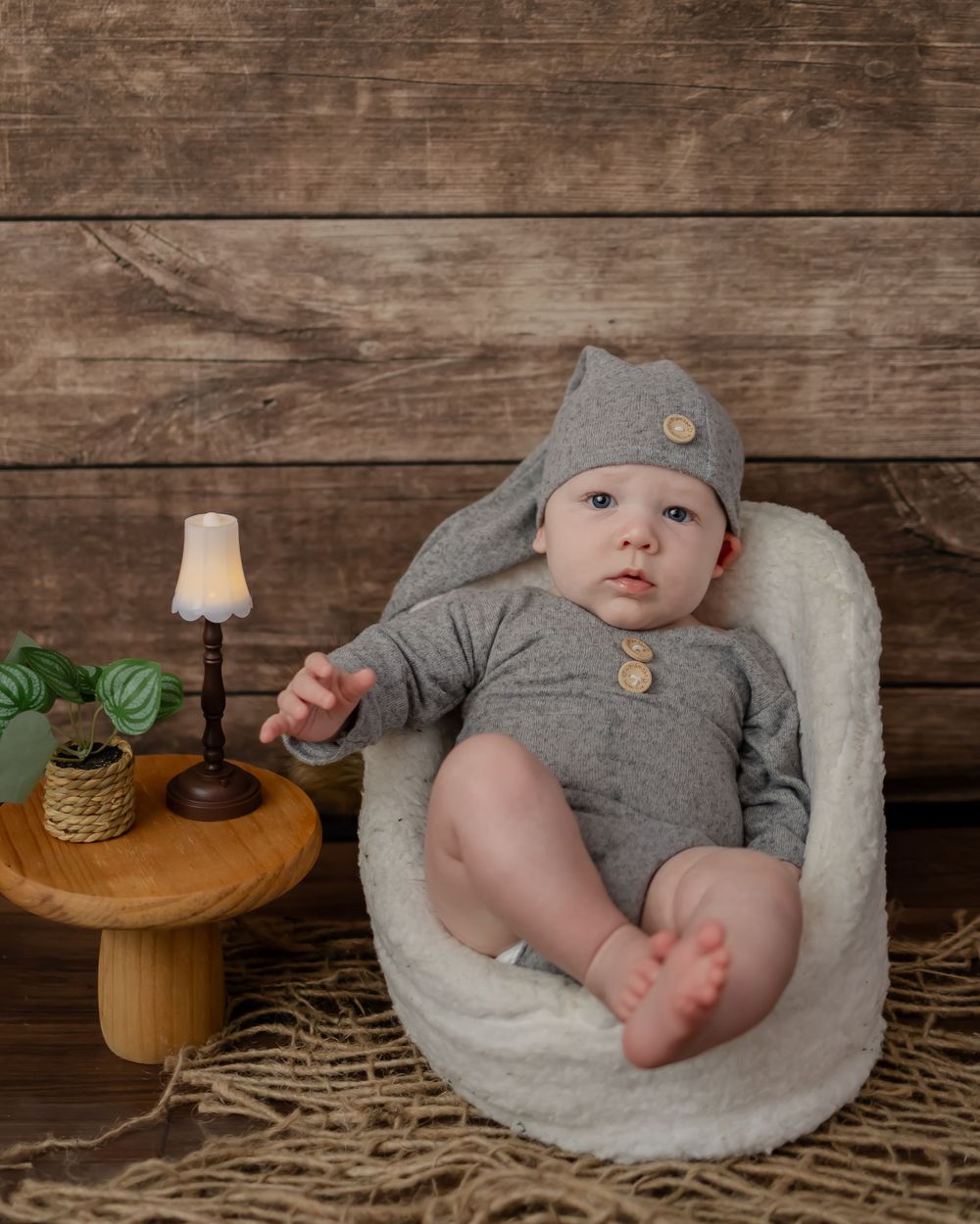 Baby in gray outfit rests in white cushion with rustic wood background and table lamp.