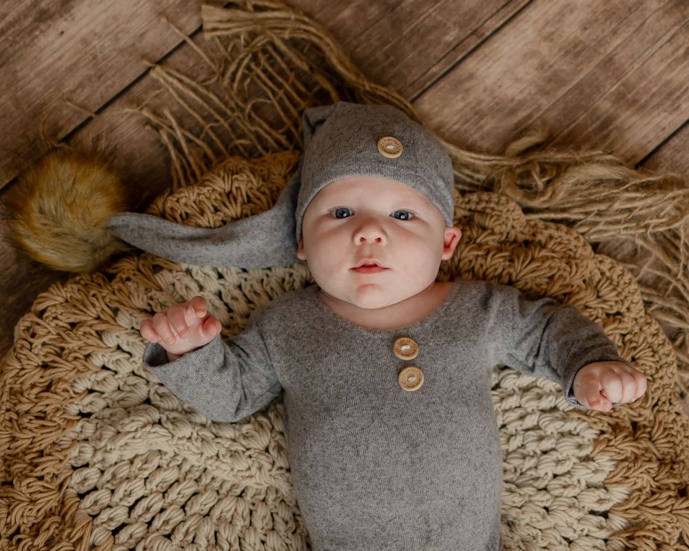 Baby wearing gray buttoned outfit and cap lying on knitted blanket against wooden backdrop.