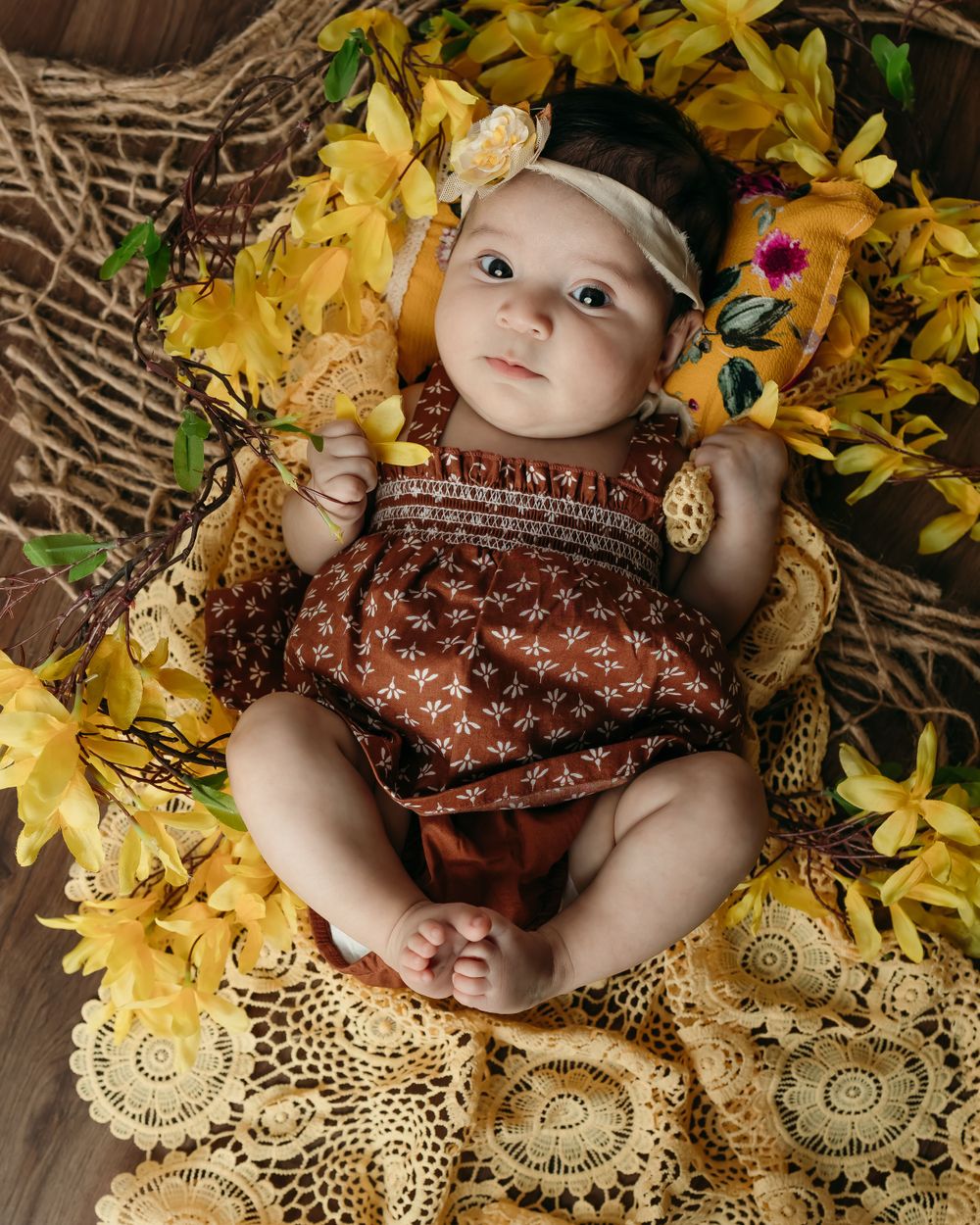 Baby in brown dress posed with yellow flowers and vintage doily decorations.