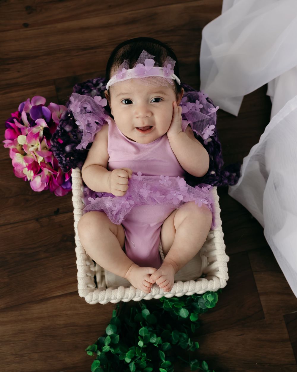 Baby in pink tulle outfit with floral accessories on white cushion.