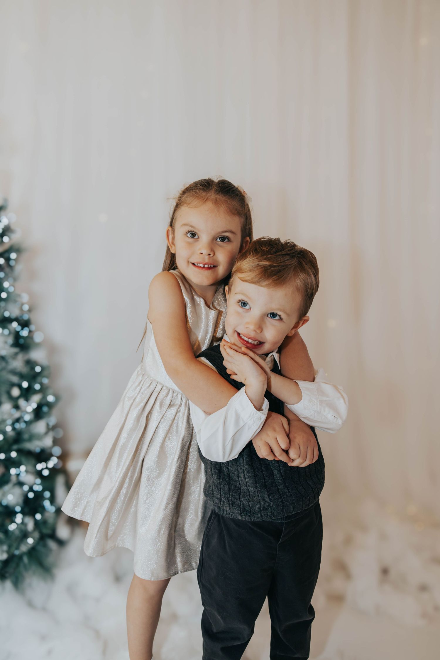 Two children in formal attire pose for holiday photos beside a decorated Christmas tree.
