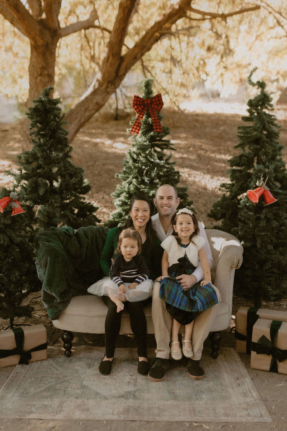 Family sits on beige armchair between decorated Christmas trees with red bows in outdoor holiday setting.