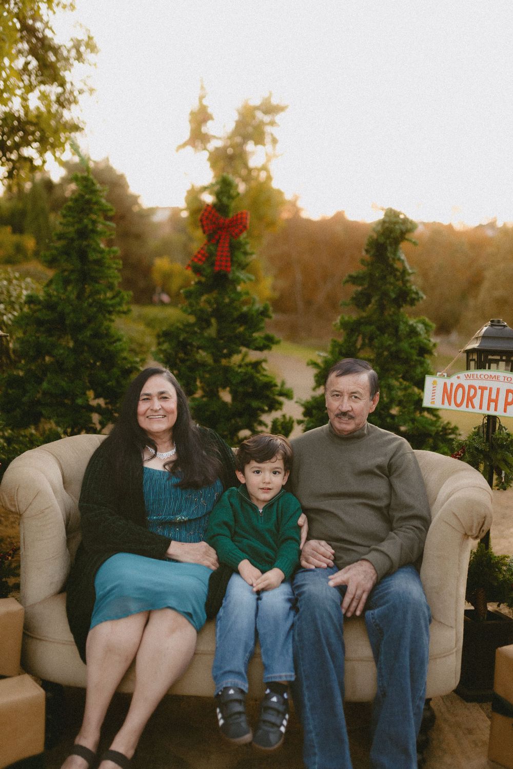 A family sits together on a beige couch outdoors with Christmas decorations and evergreen trees in the background at sunset.