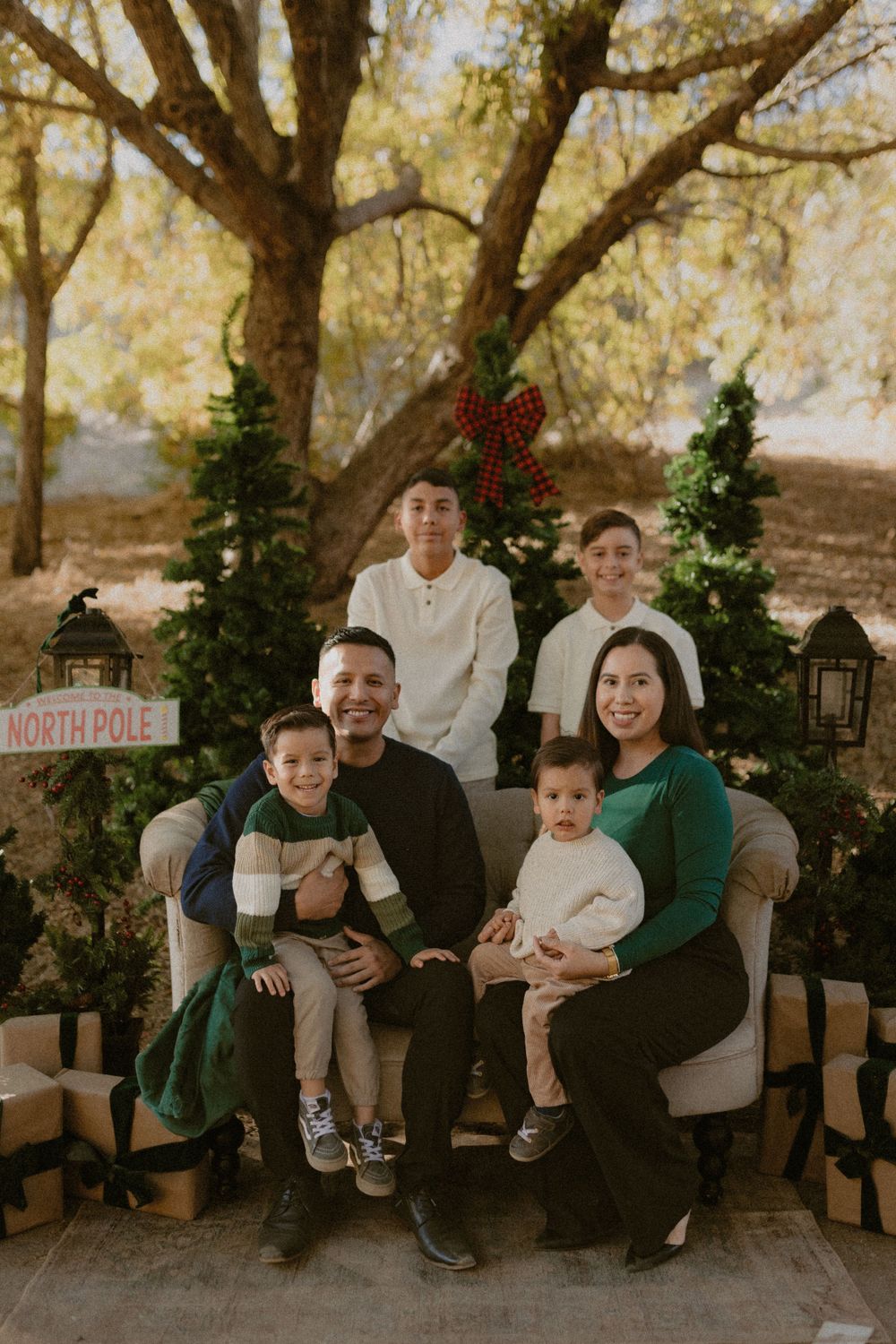 A family sits together on a bench with Christmas decor and trees in the background during a holiday photoshoot.