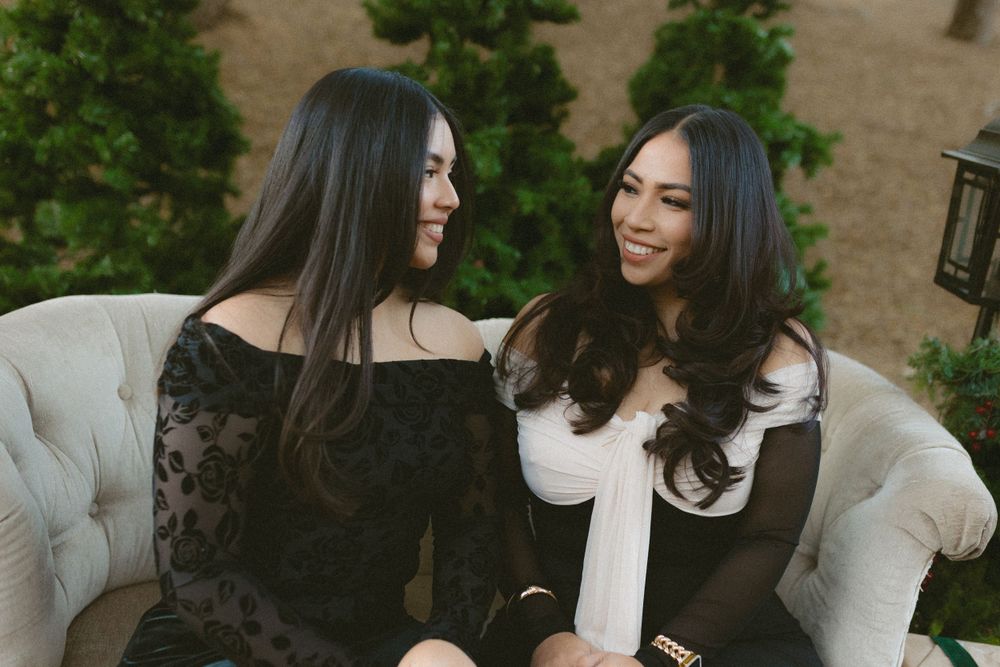 Two women in black and white outfits sit together on a light gray couch while smiling and looking at each other.