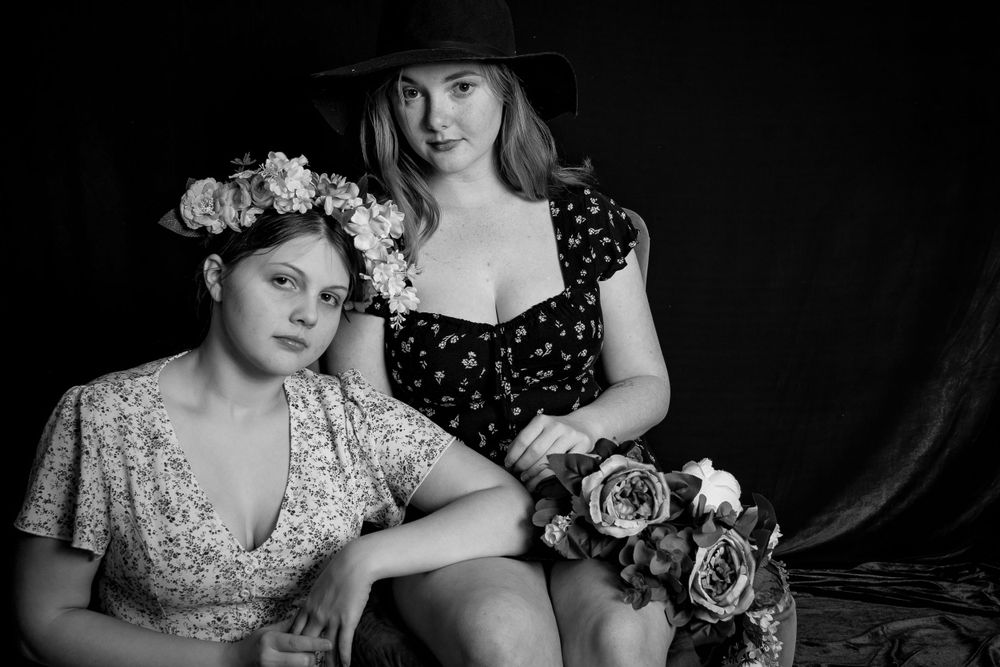 A black and white artistic portrait of two people wearing floral dresses and flower crowns against a dark backdrop.