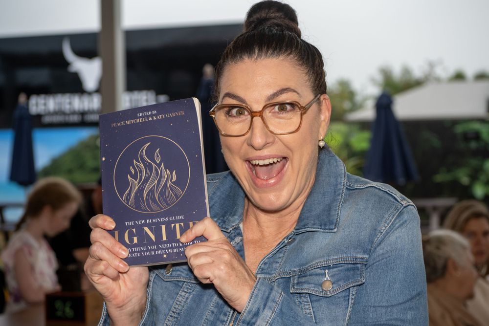 Someone in glasses and a denim jacket excitedly holds up a blue  book outdoors.