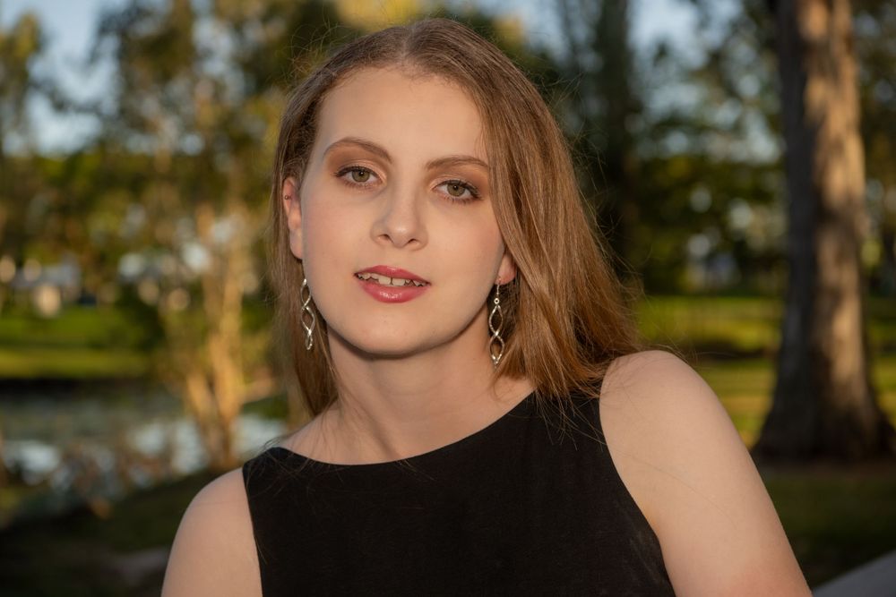 A woman in a black sleeveless top poses outdoors at dusk with trees and water in the background.