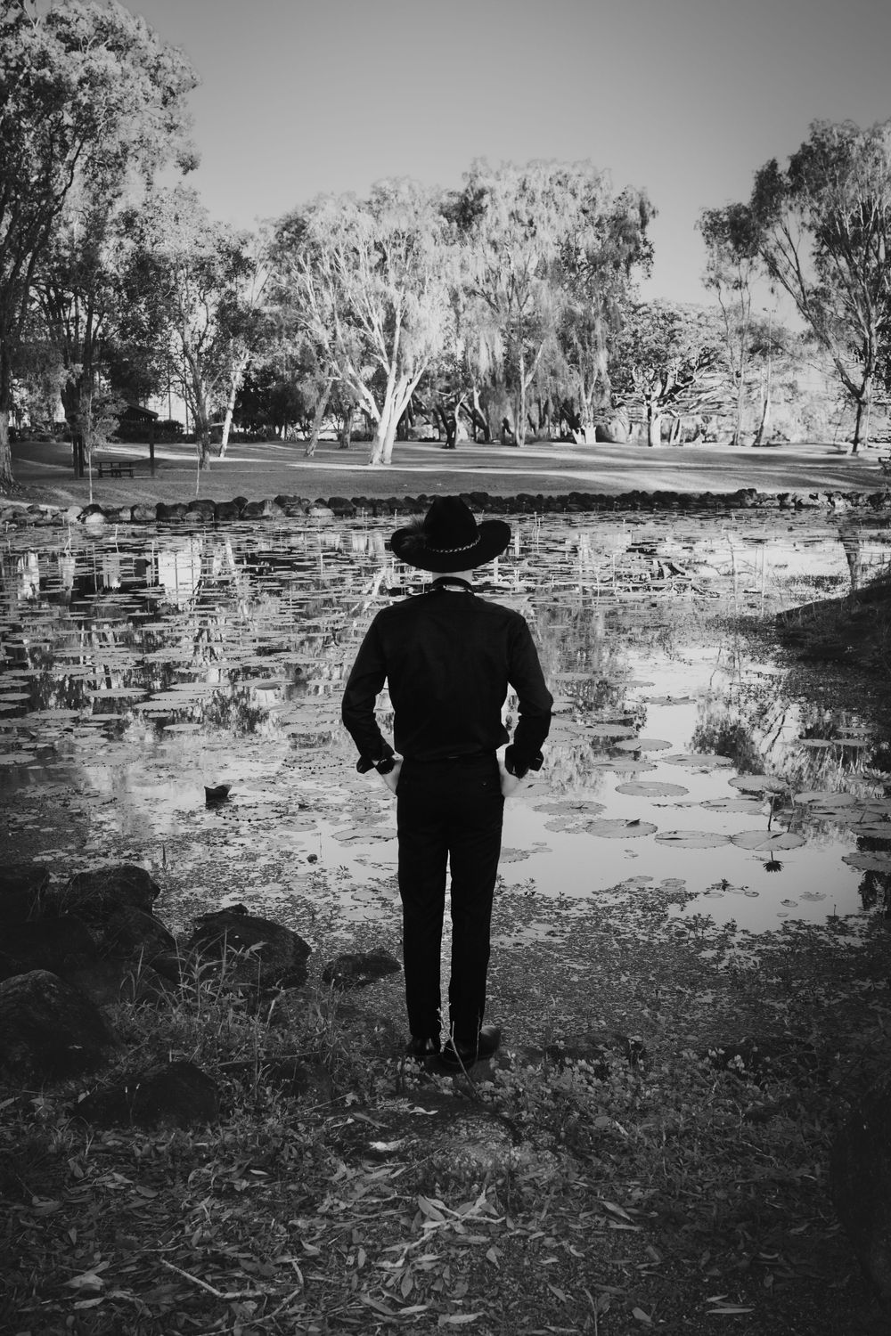 A silhouetted figure in a hat stands facing a reflective pond with eucalyptus trees in the background, shot in black and white.