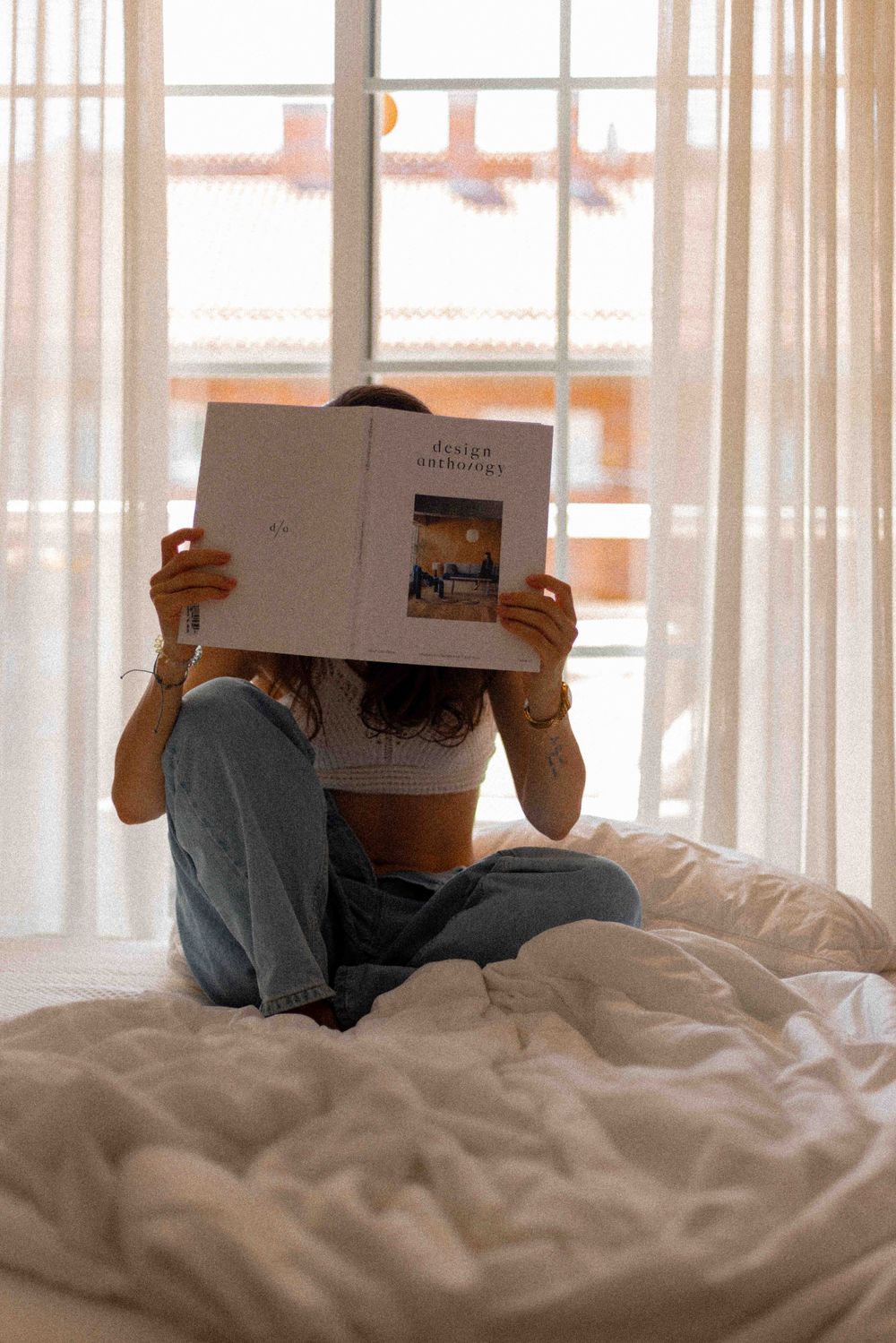 Person reading book while relaxing on white bedding near sunlit window.