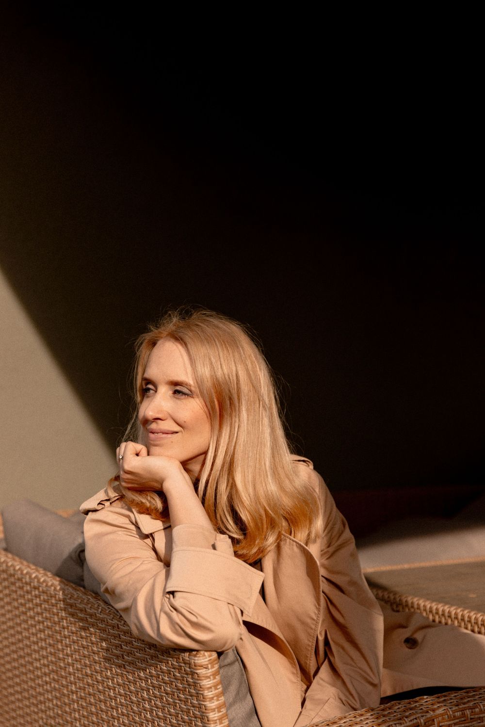 Woman in beige jacket sits on woven outdoor furniture while resting her head on hand in warm evening lighting.