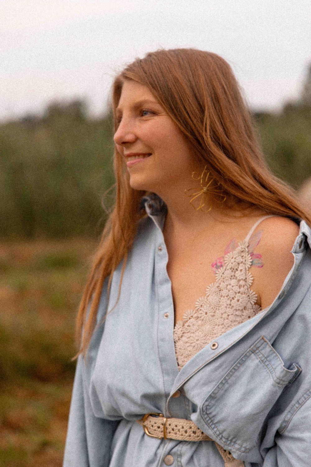 A person with long brown hair wearing a denim jacket and lace top looks to the side while outdoors at sunset.