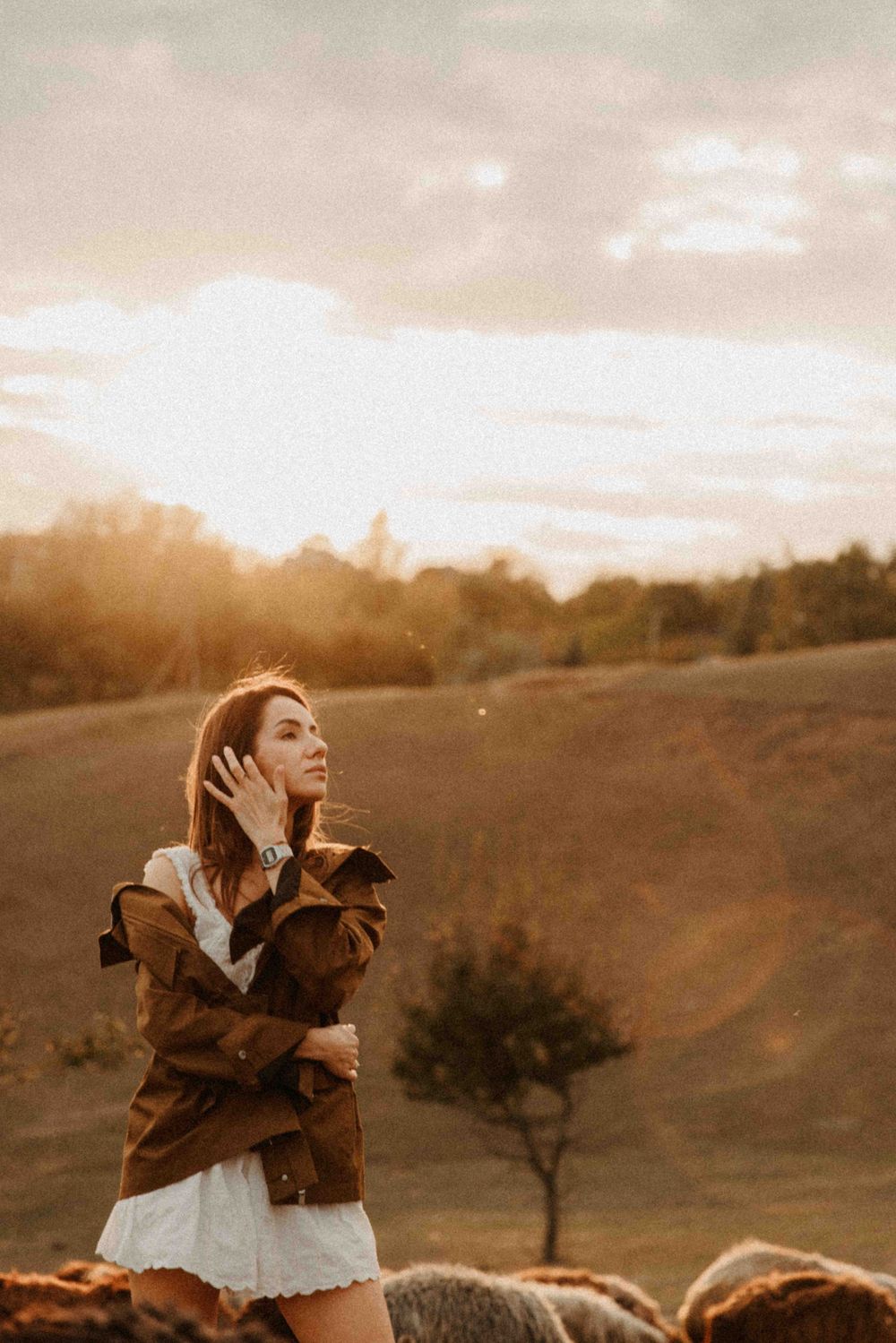 A dreamy sunset scene in a countryside field with a white dress and brown jacket against a warm golden sky.