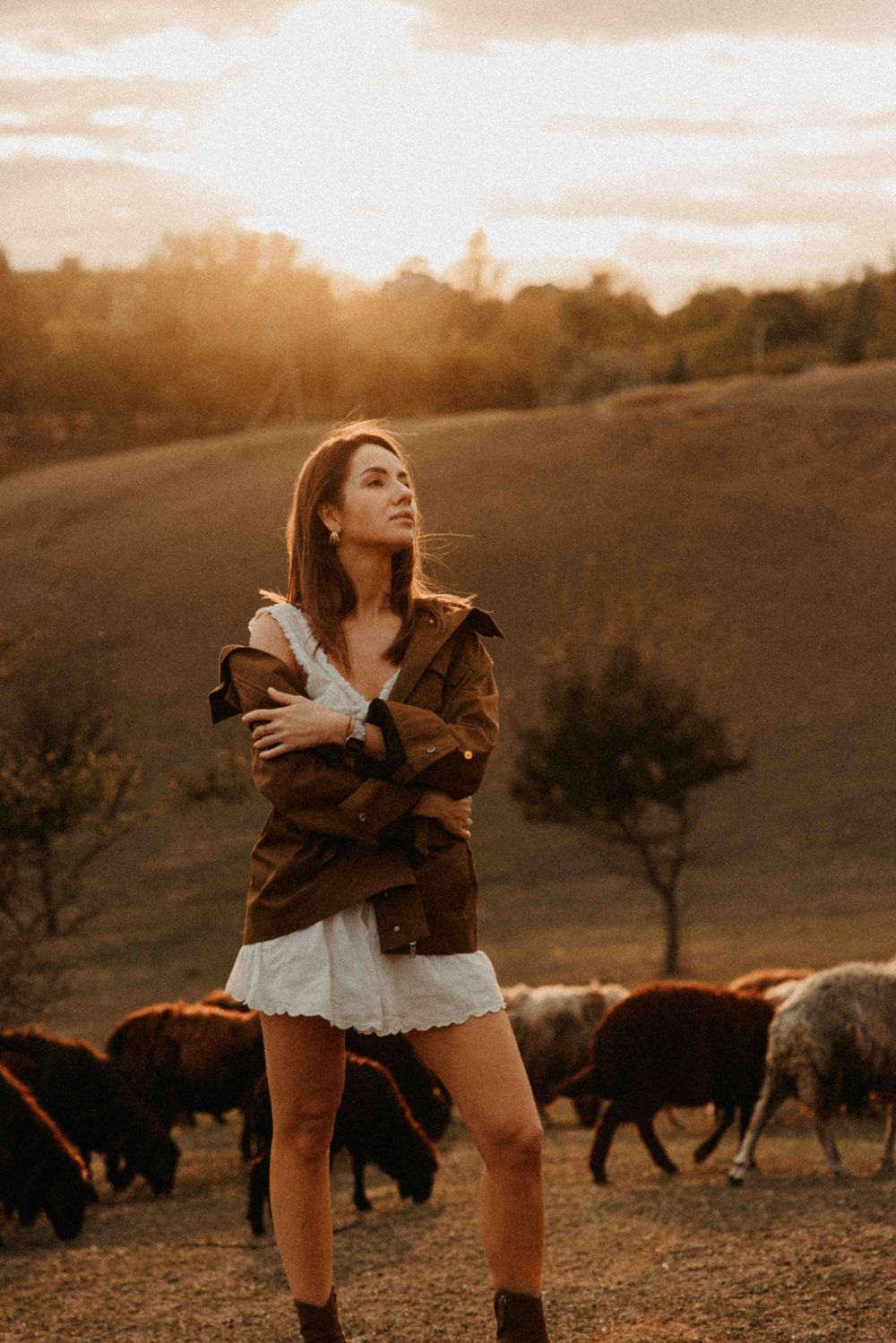 Someone in a brown jacket and white dress poses in a field at sunset while buffalo graze in the background.
