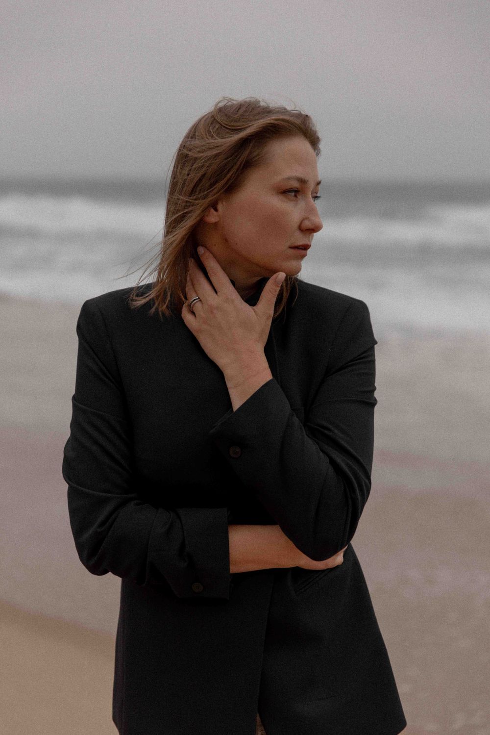 Contemplative person in black blazer stands on misty beach with windswept hair against moody grey ocean backdrop.