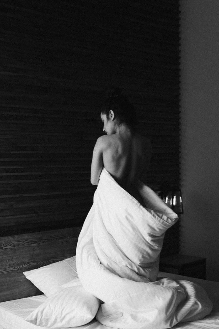 Artistic black and white photo of person wrapped in white bedding sitting on floor.