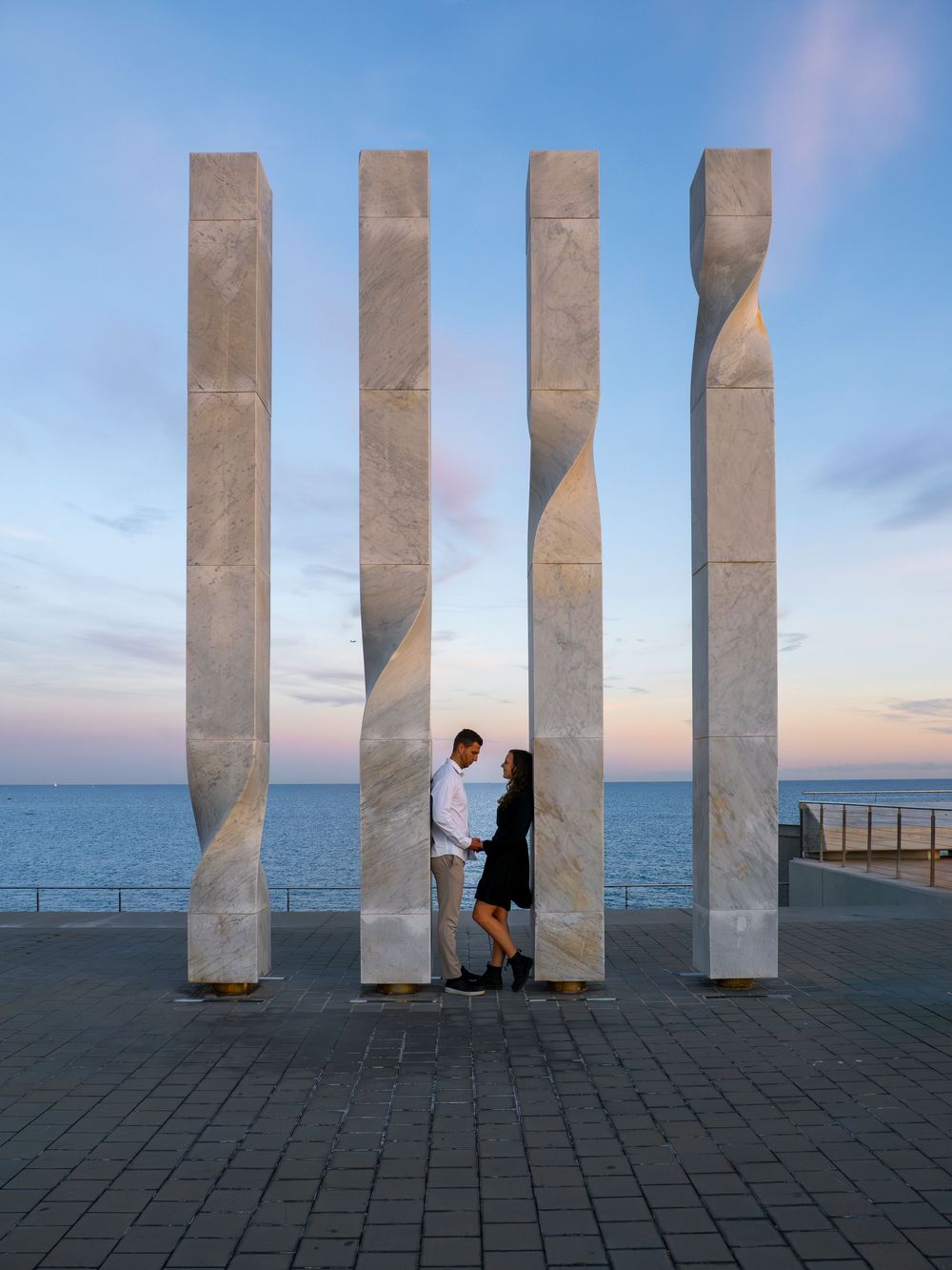 Artistic engagement photos between white concrete pillars against ocean backdrop at sunset.