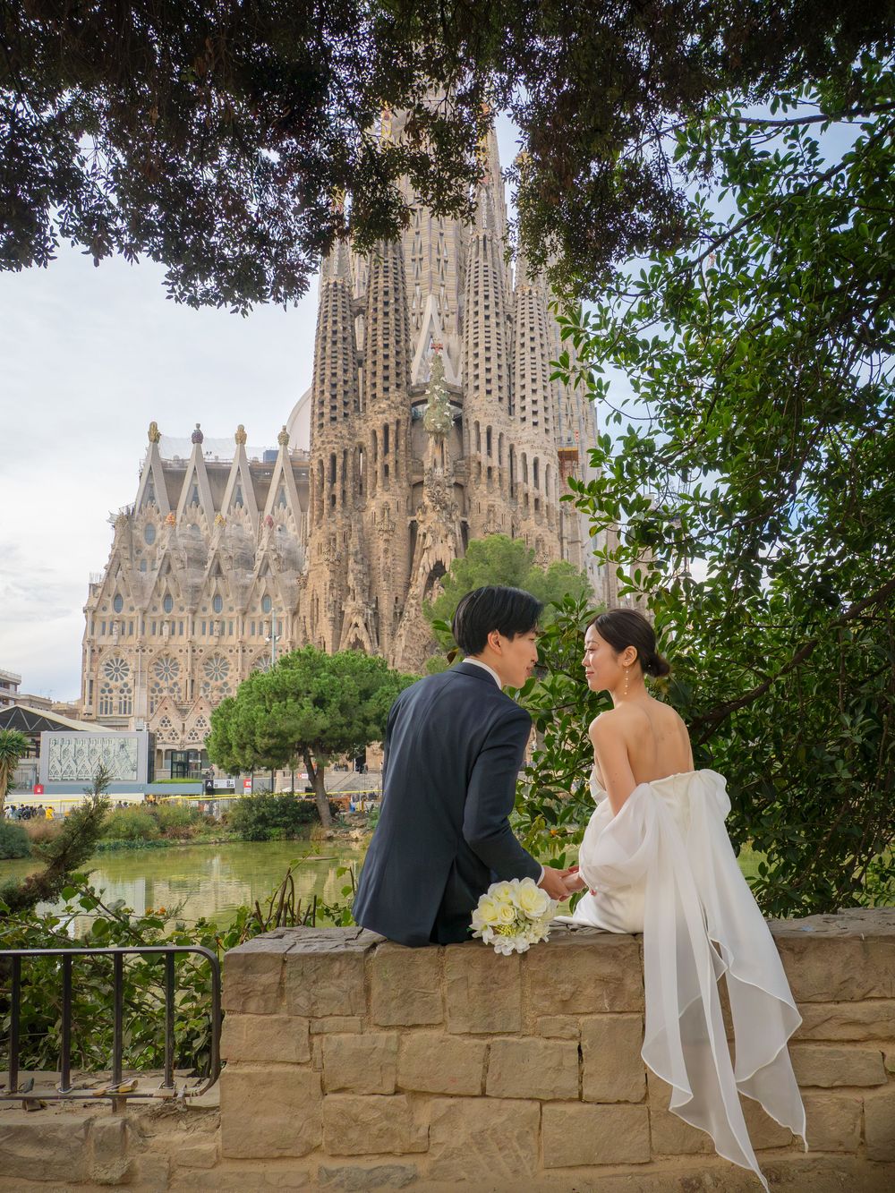 A couple poses together on a stone wall with Barcelona's Sagrada Familia cathedral in the background.