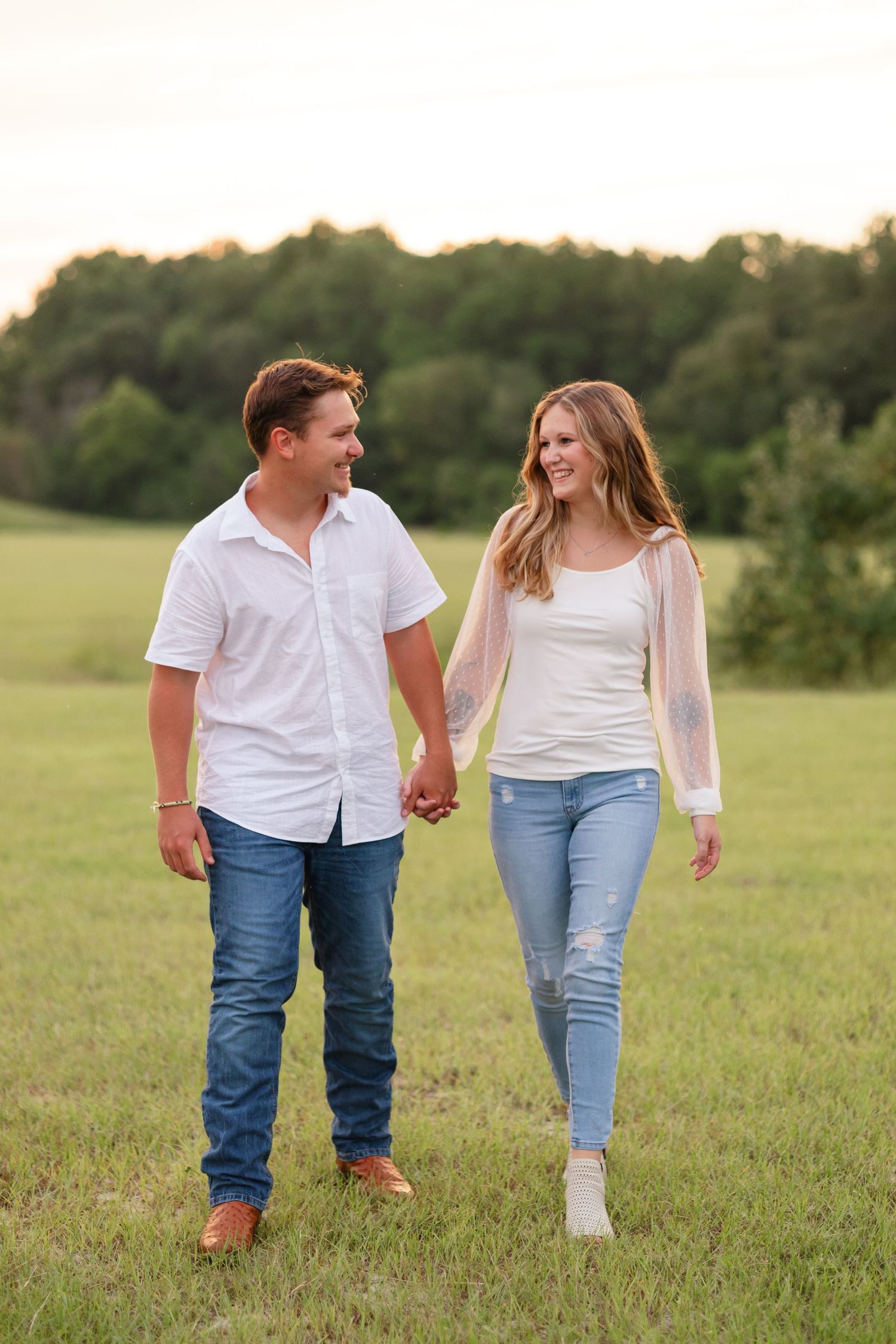 A couple holds hands while walking through a sunlit meadow in casual summer attire.