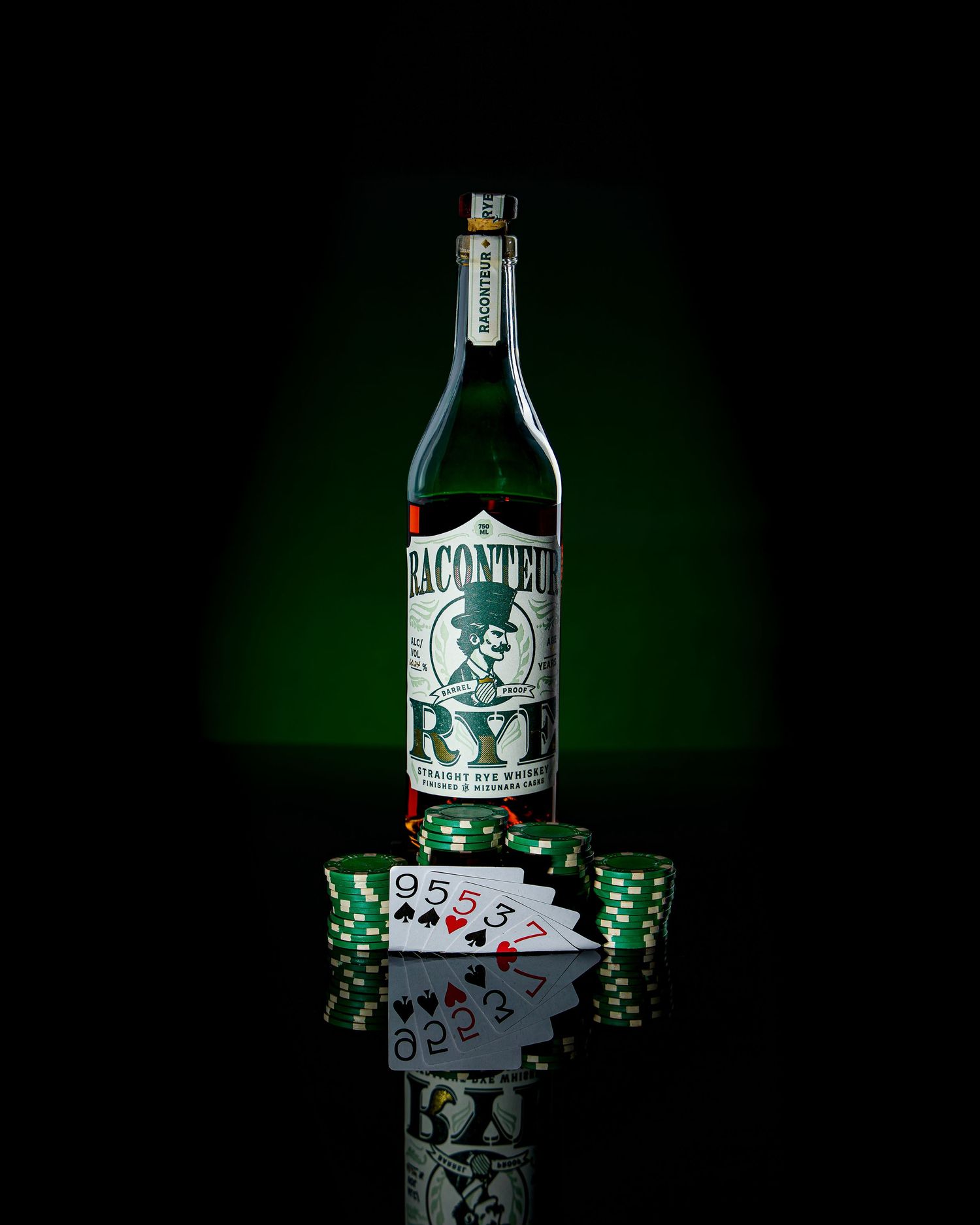 Bottle of Raconteur Rye with playing cards and poker chips reflected on glossy black surface in green lighting.