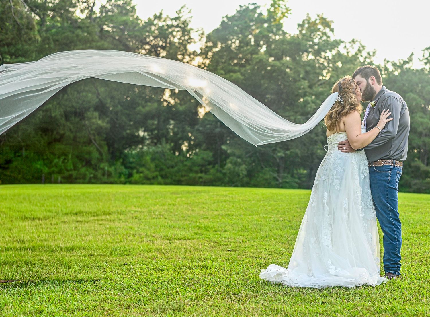 A bride in a flowing white gown with veil billowing in the wind embraces her groom on a lush green lawn.