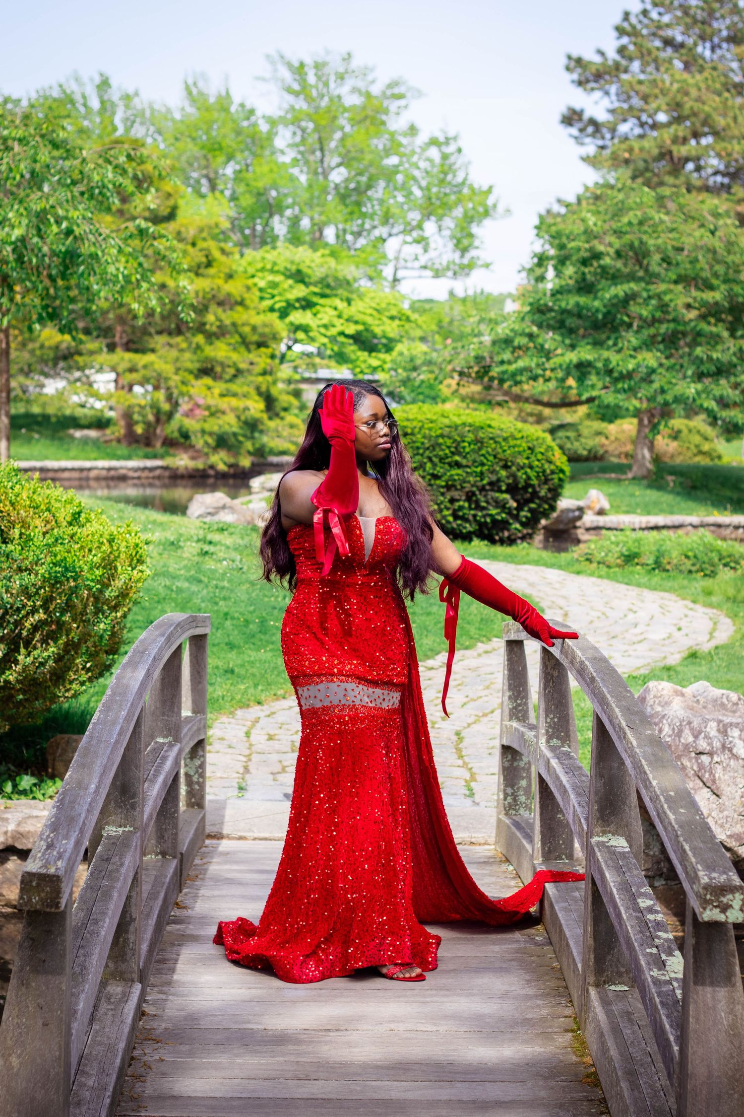 Person in sparkling red formal dress striking dramatic pose on wooden footbridge in garden setting.