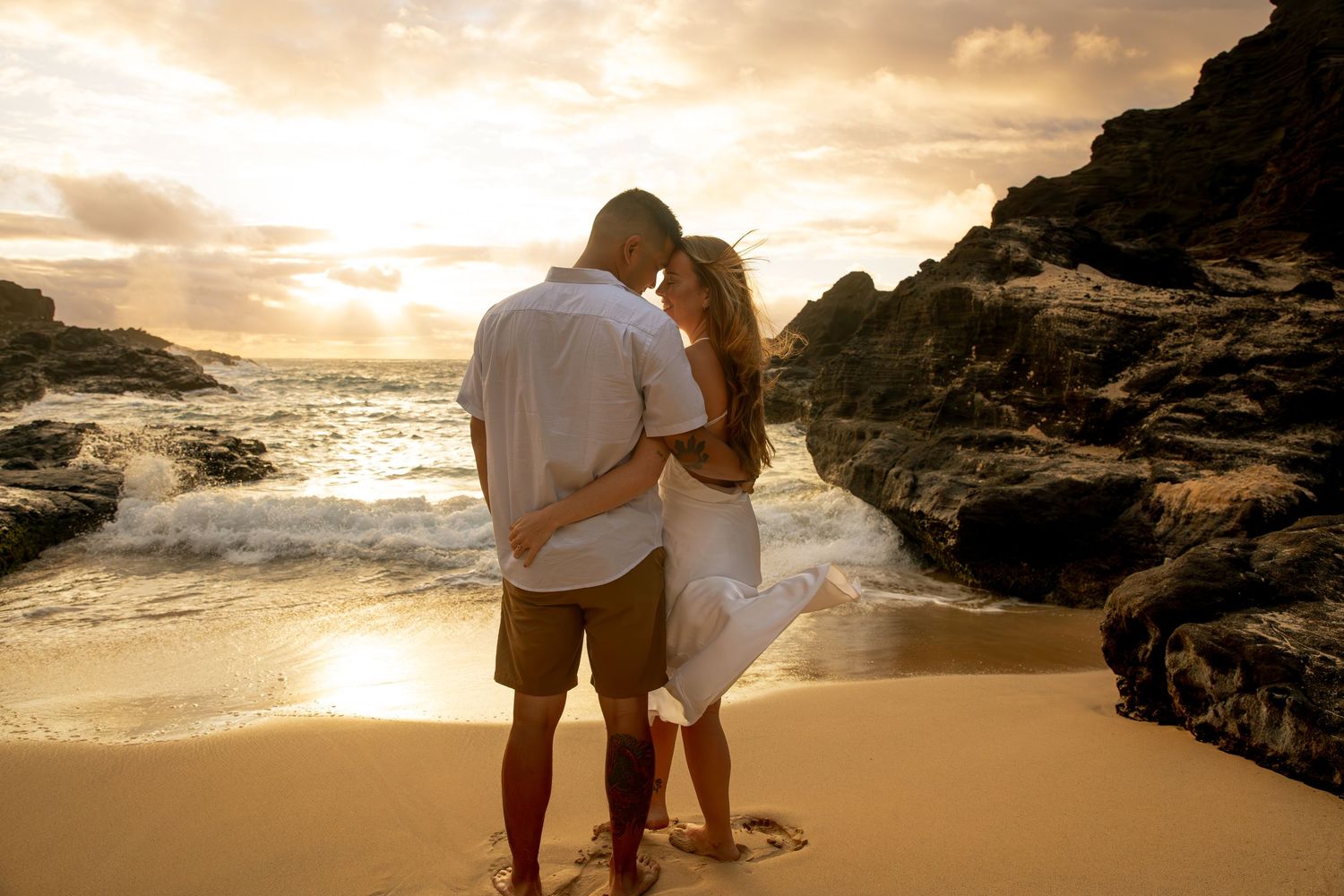 A couple shares a romantic moment on a beach at sunset with waves crashing against rocky cliffs in Hawaii.