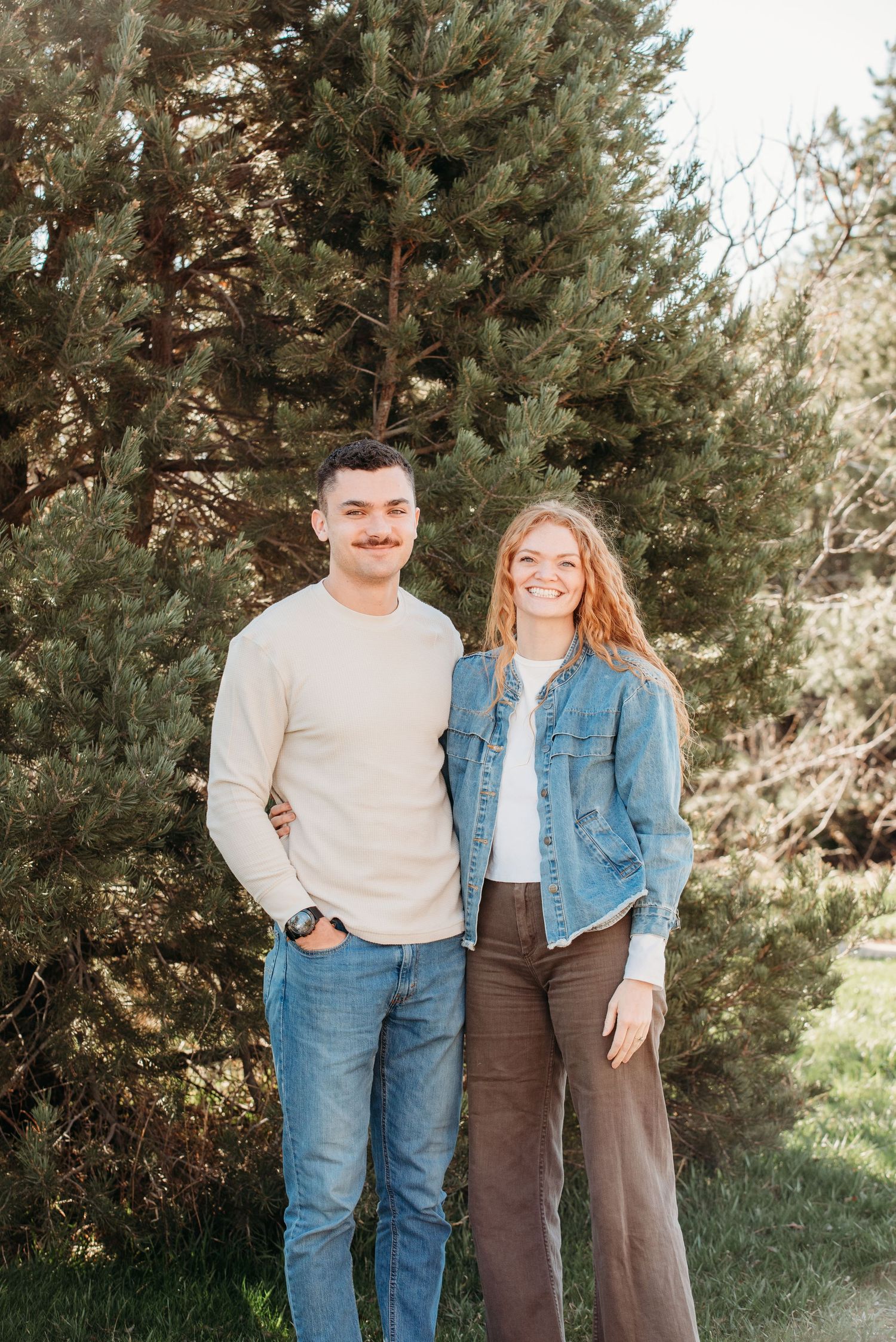 A couple poses casually in front of an evergreen tree wearing coordinated casual outfits in a natural outdoor setting.