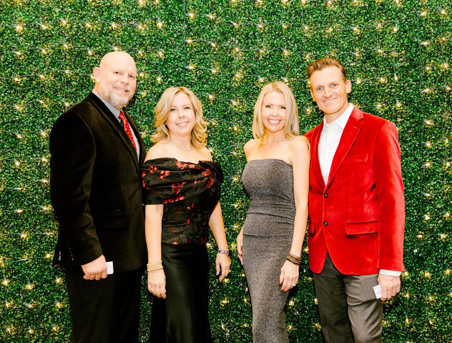 Two couples pose together against a green hedge wall backdrop at a formal event, wearing elegant evening attire.
