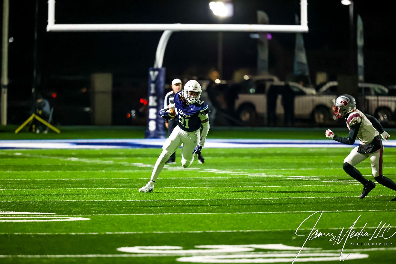 Football players run downfield during a nighttime game on an illuminated artificial turf field.