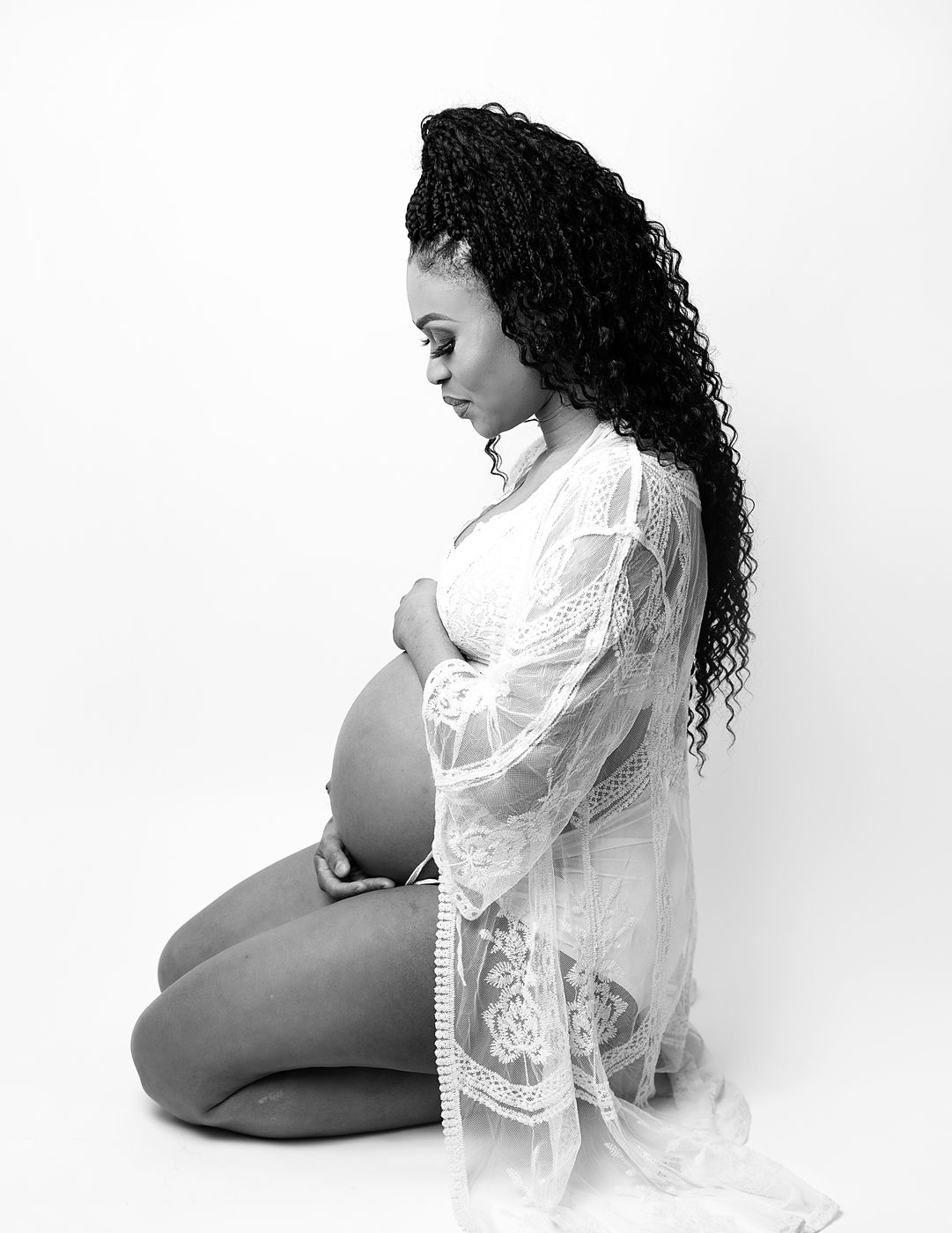A black and white maternity photo in London shows an expectant mother wearing a white lace robe while kneeling gracefully in profile.