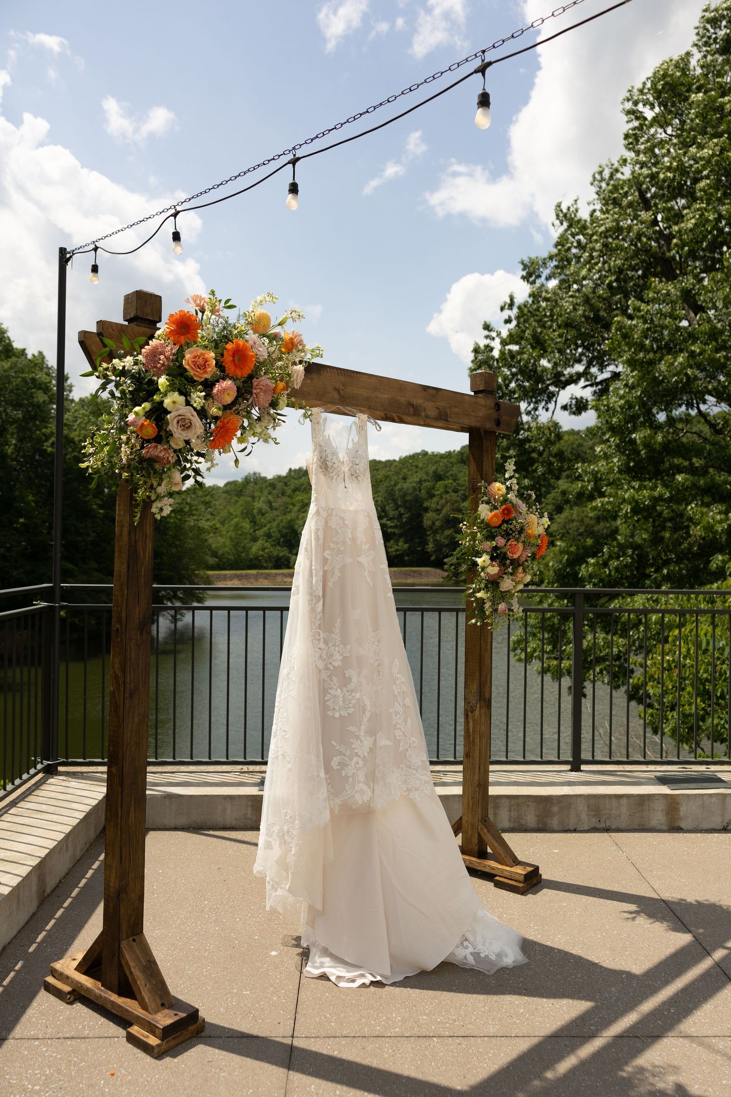 White wedding dress hangs from wooden arch with string lights and floral decorations by a lake.