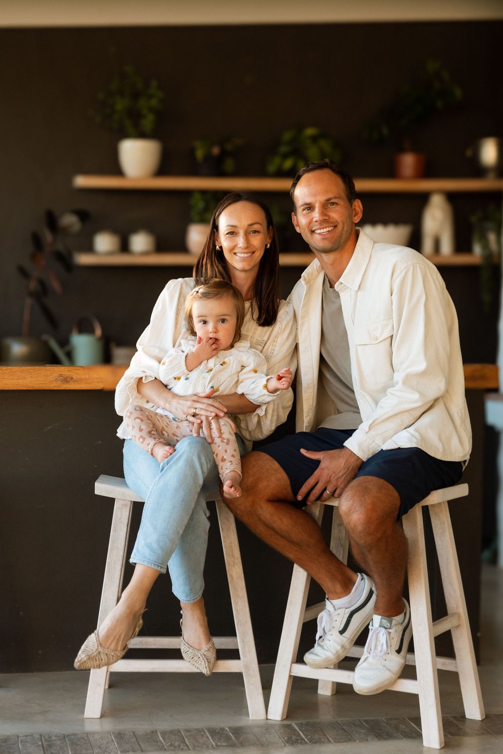 Family sitting in white chairs against dark wall with decorative floating shelves and potted plants.