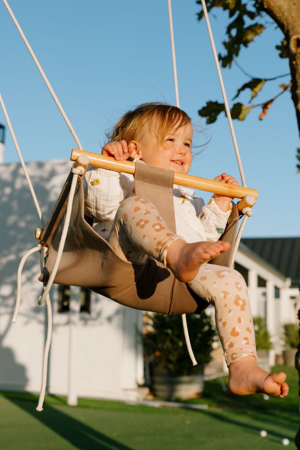 A joyful toddler swings outdoors in a brown canvas swing against a blue sky background.