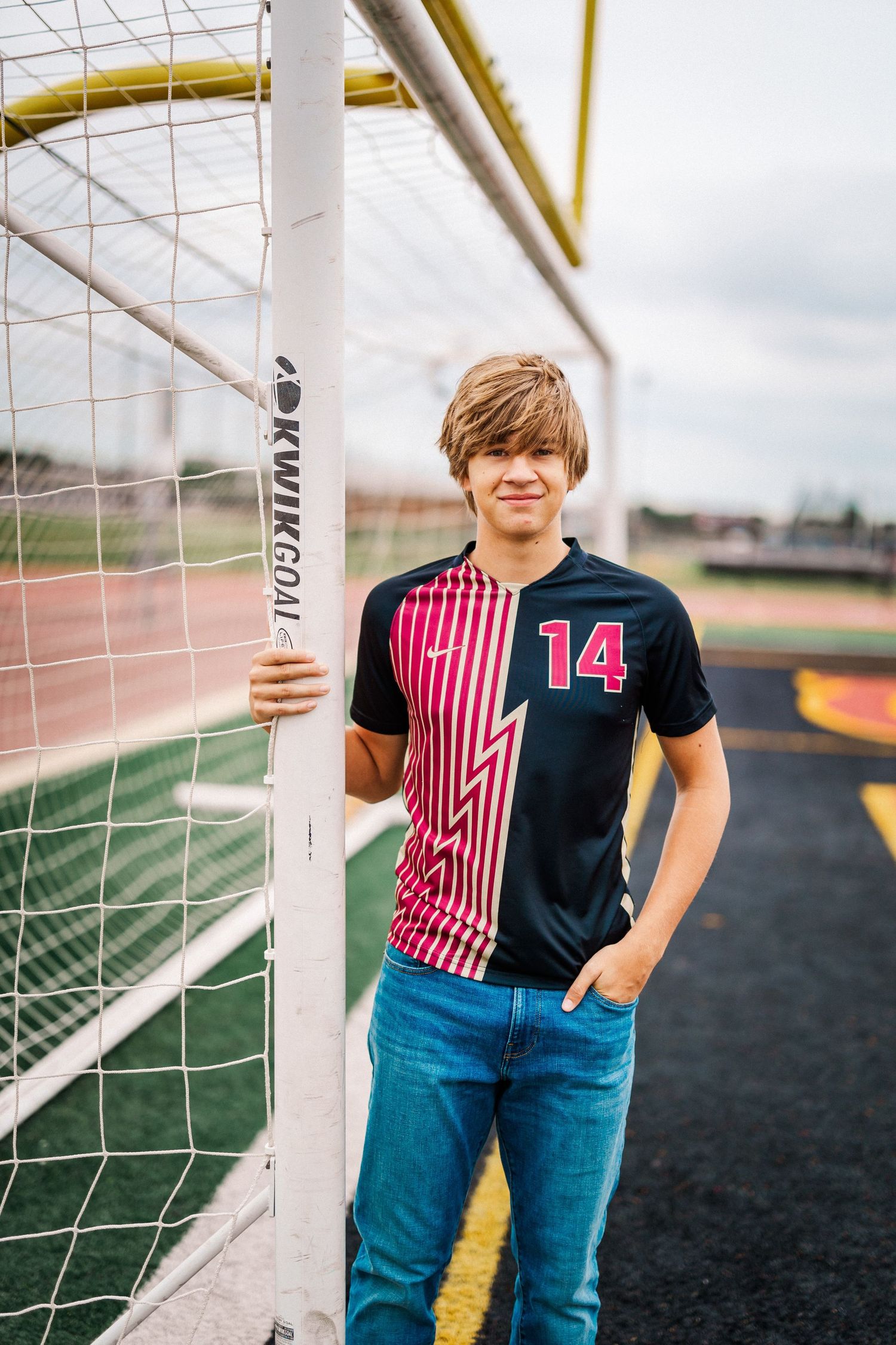 Teen boy in soccer jersey number 14 leaning against goal post on athletic field.