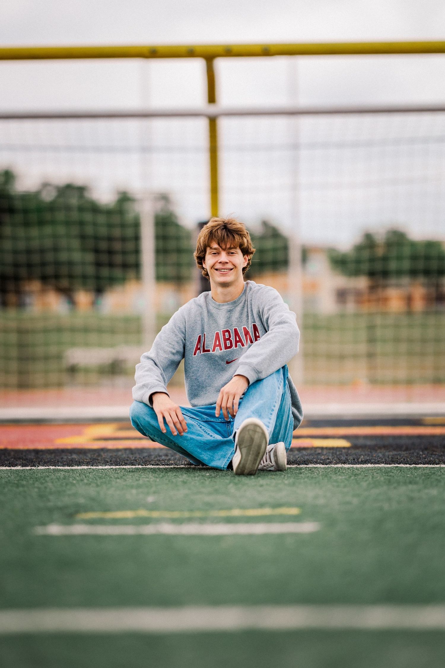 Teen in Alabama sweatshirt sitting cross-legged on green turf field smiling at camera.