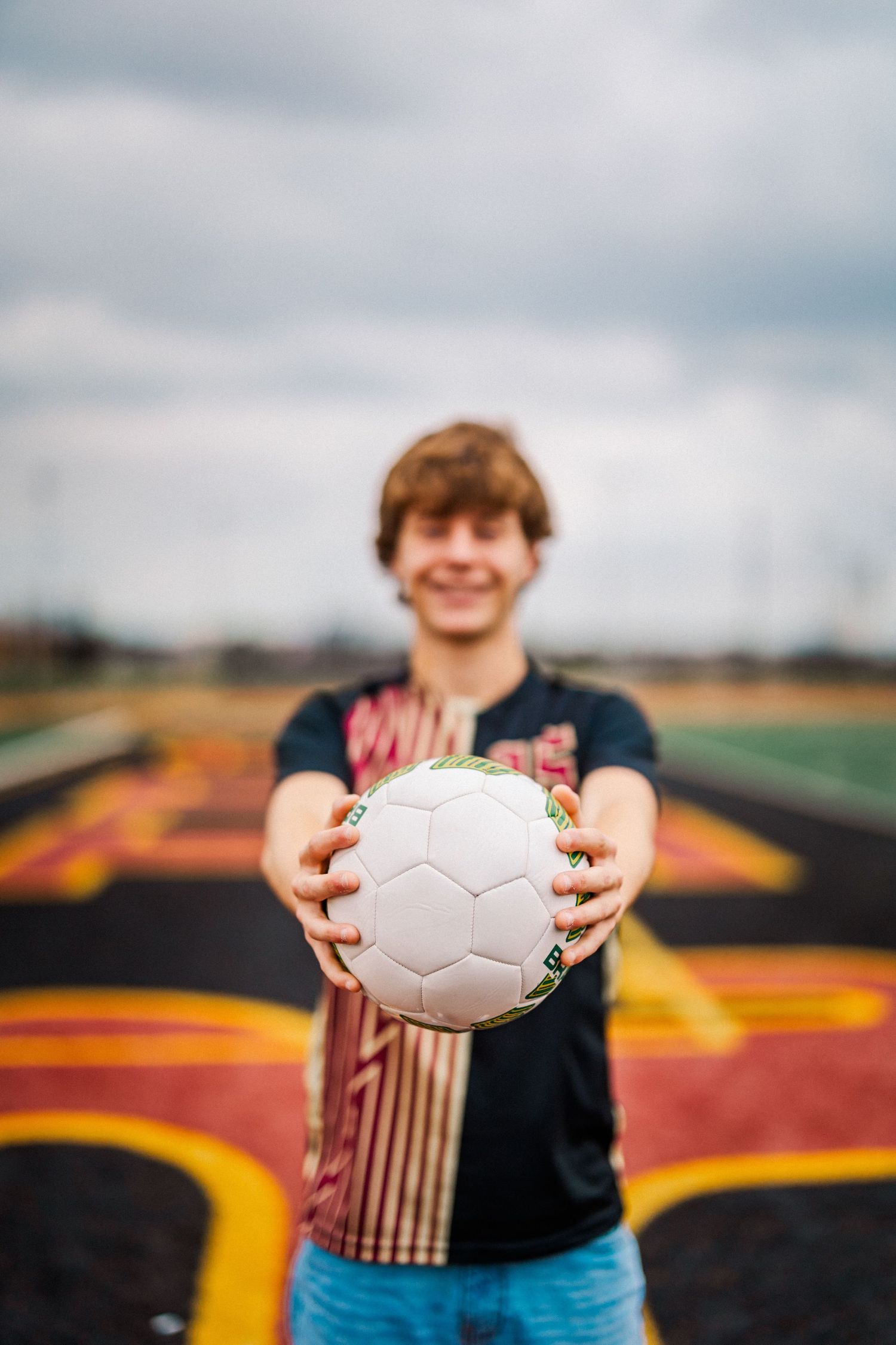 Smiling teen holding a soccer ball toward the camera on a colorful athletic track.