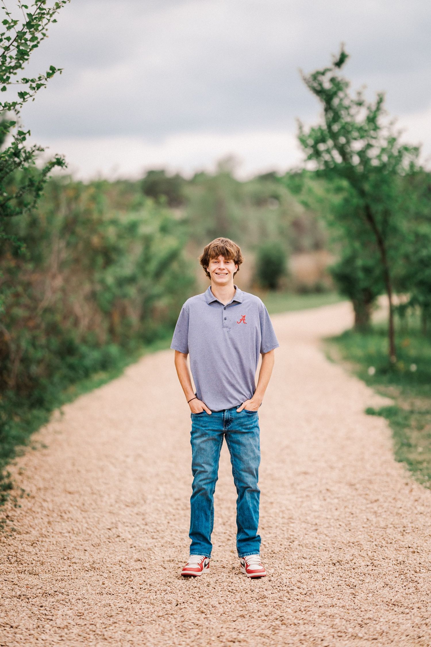 Smiling teen boy in a polo shirt standing on a gravel path lined with green trees.