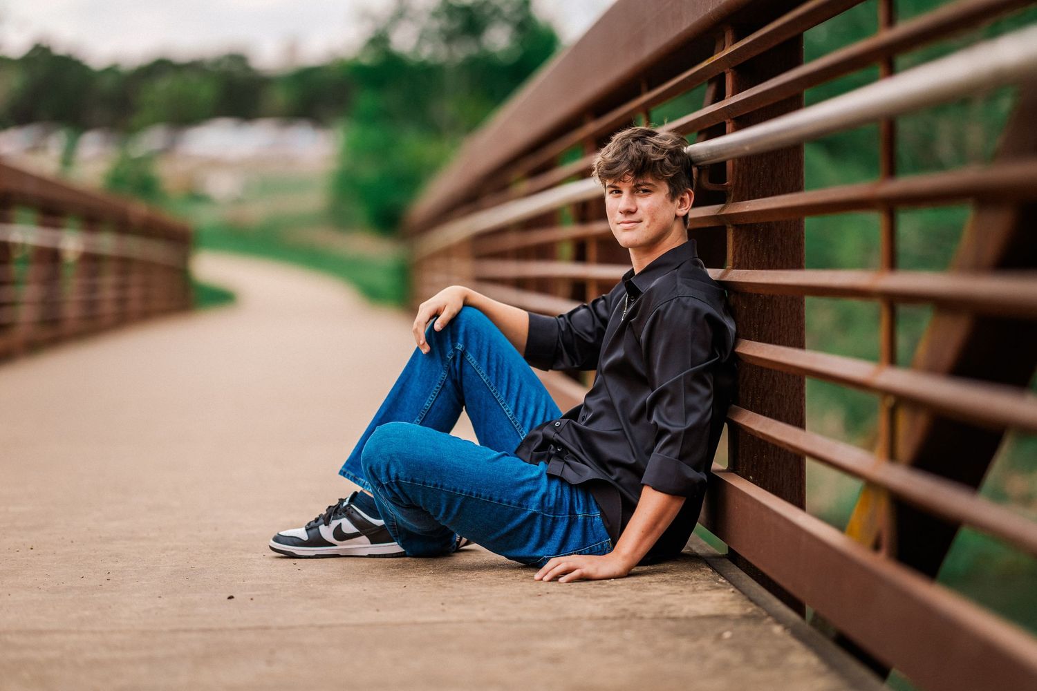 Teen boy in a black shirt sitting relaxed on a bridge railing with blue jeans.