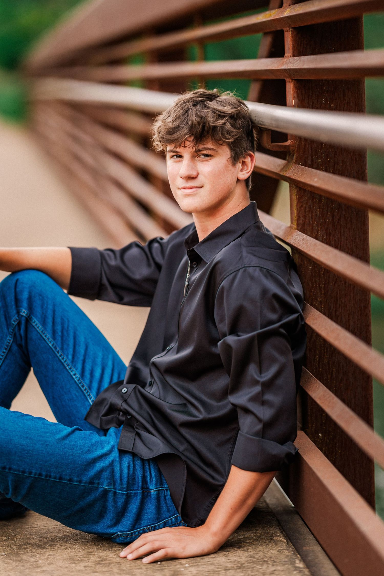 Teen boy in black shirt and jeans sits relaxed against a rustic metal bridge railing outdoors.
