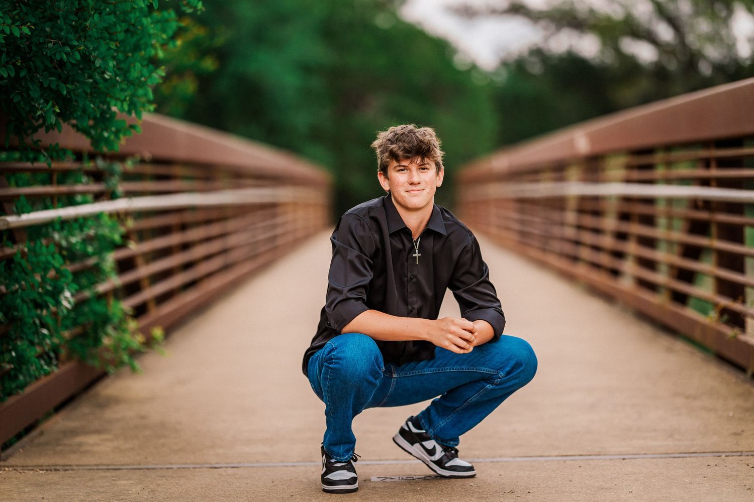 Teen boy crouching confidently on a wooden bridge surrounded by lush green foliage.