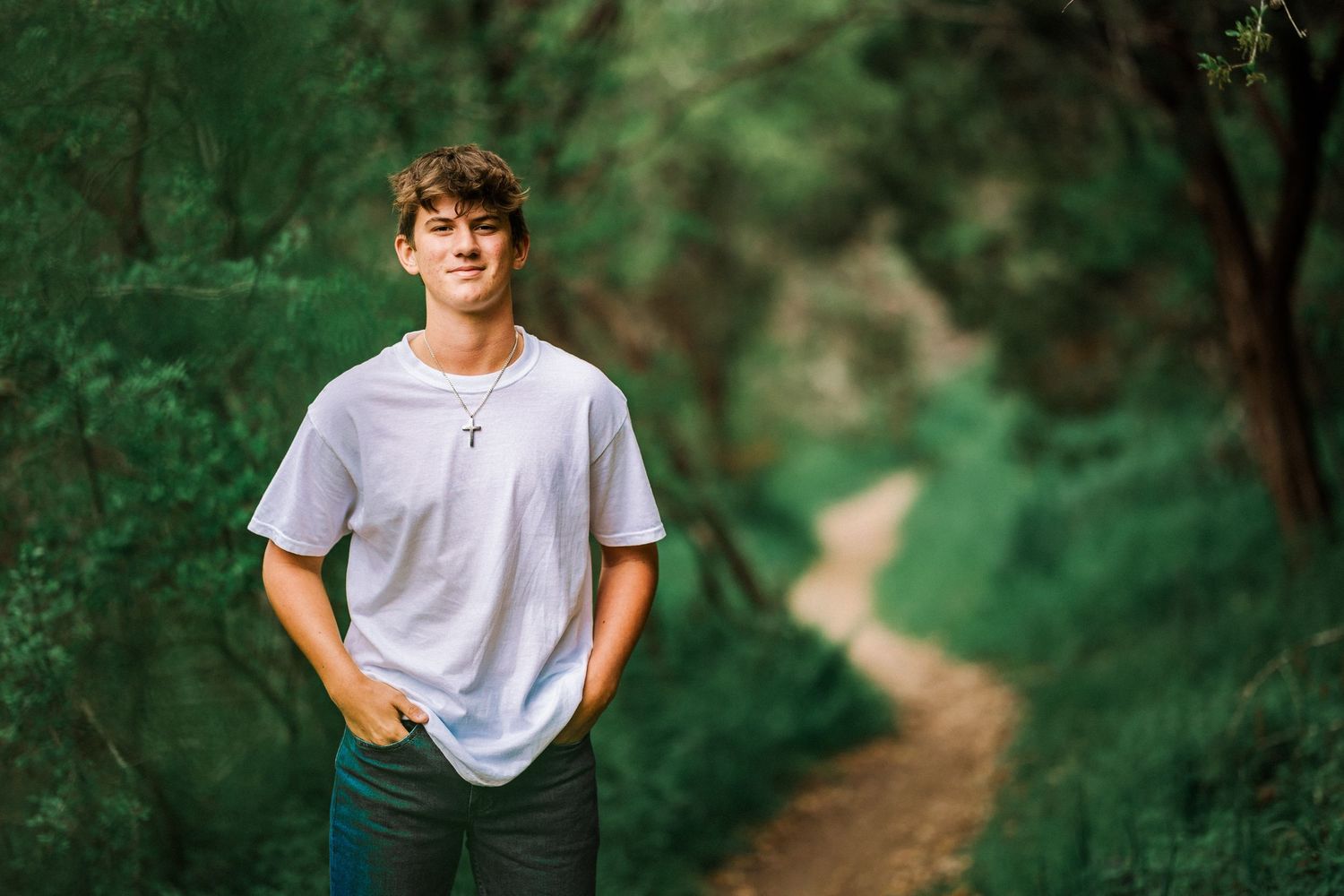 Young man in a white t-shirt and cross necklace standing on a wooded trail path.