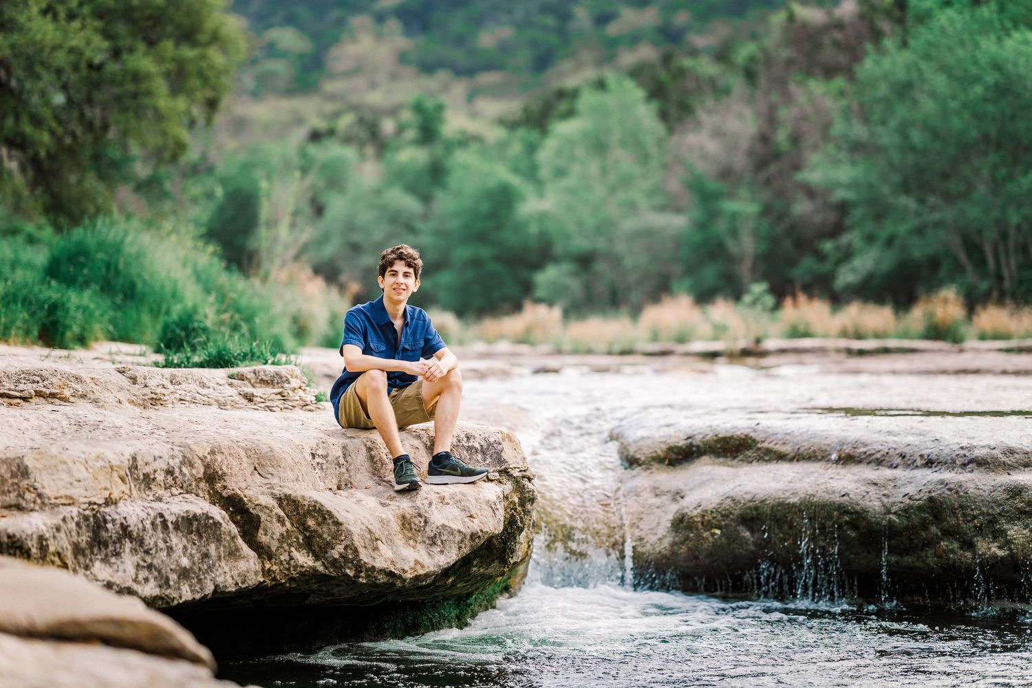 Young man sitting on rocks by a flowing stream surrounded by lush green trees in nature.