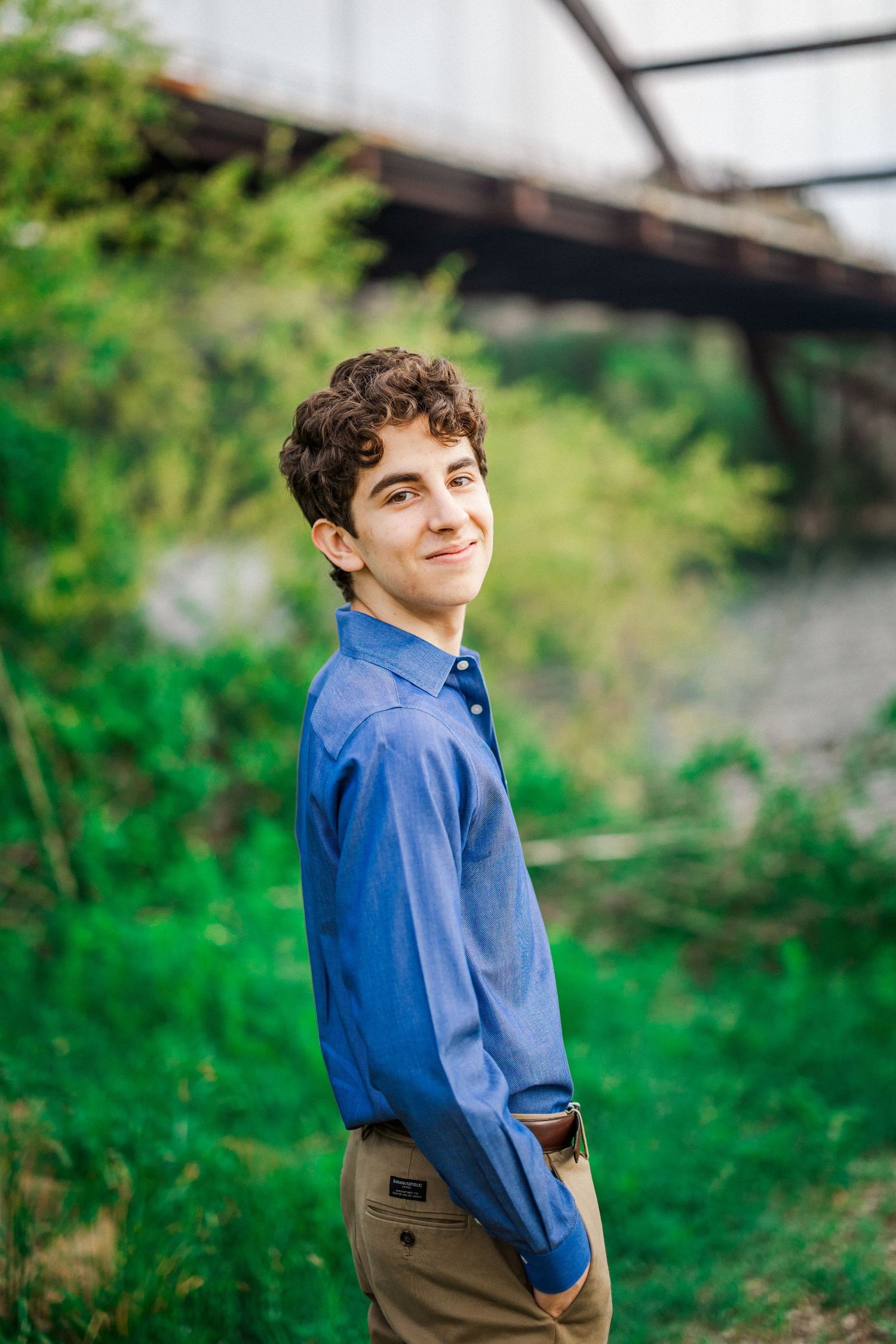 Smiling young man in a blue shirt standing outdoors near a bridge and green plants.