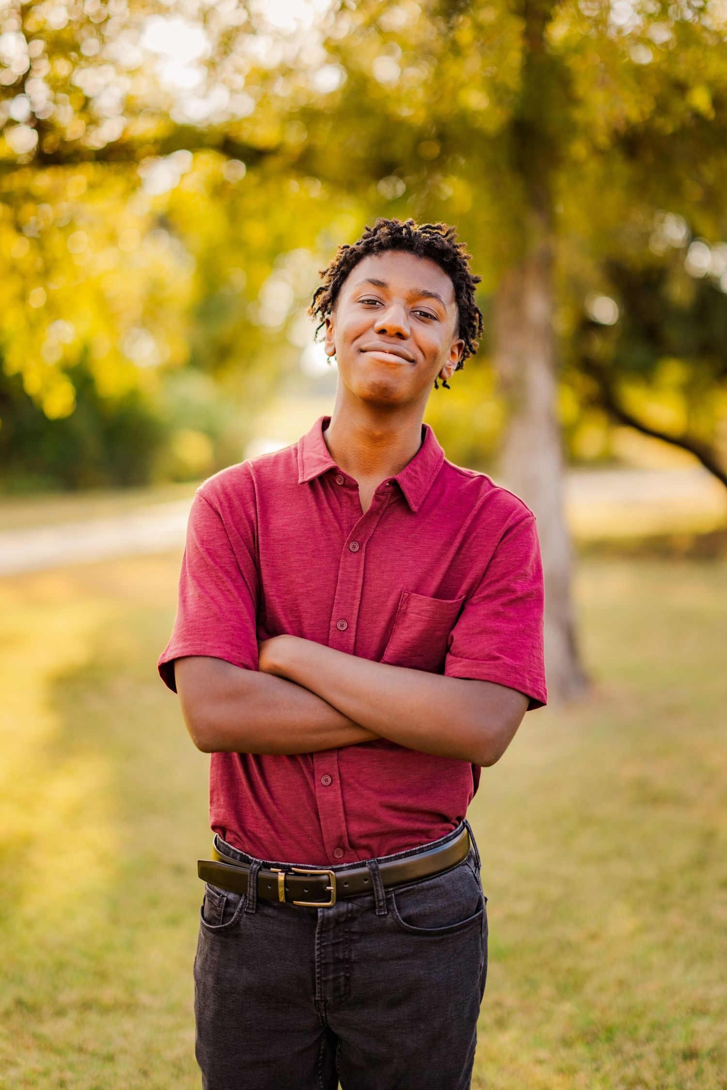 Smiling young Black man in red shirt standing with arms crossed in a sunny park setting.